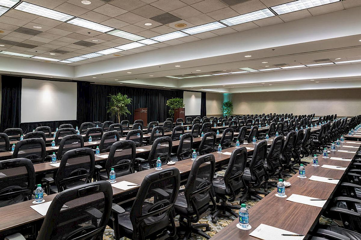 A large conference or meeting room with many chairs, desks, and water bottles, set up for an event or seminar. There are two screens at the front.