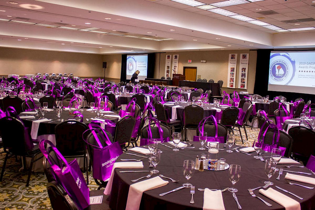 A conference hall with round tables set up for dining, each with purple bags on chairs, and large screens displaying logos and text.