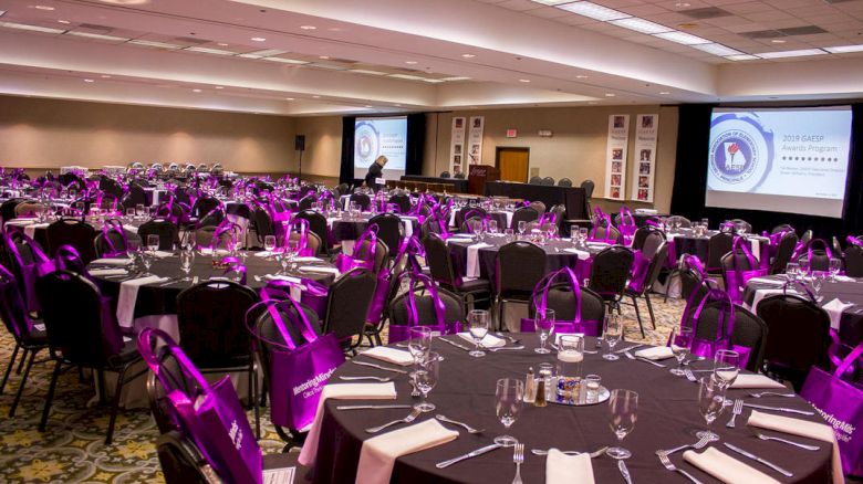 A conference hall with round tables set up for dining, each with purple bags on chairs, and large screens displaying logos and text.