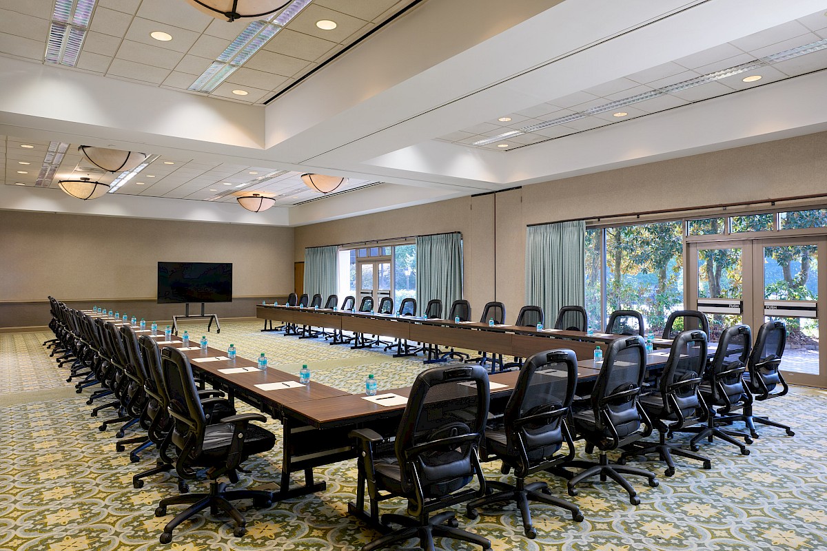 A large, empty conference room with a U-shaped table, many black rolling chairs, a wall-mounted TV, and windows with curtains.