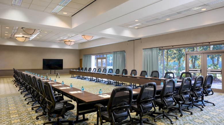 A large, empty conference room with a U-shaped table, many black rolling chairs, a wall-mounted TV, and windows with curtains.