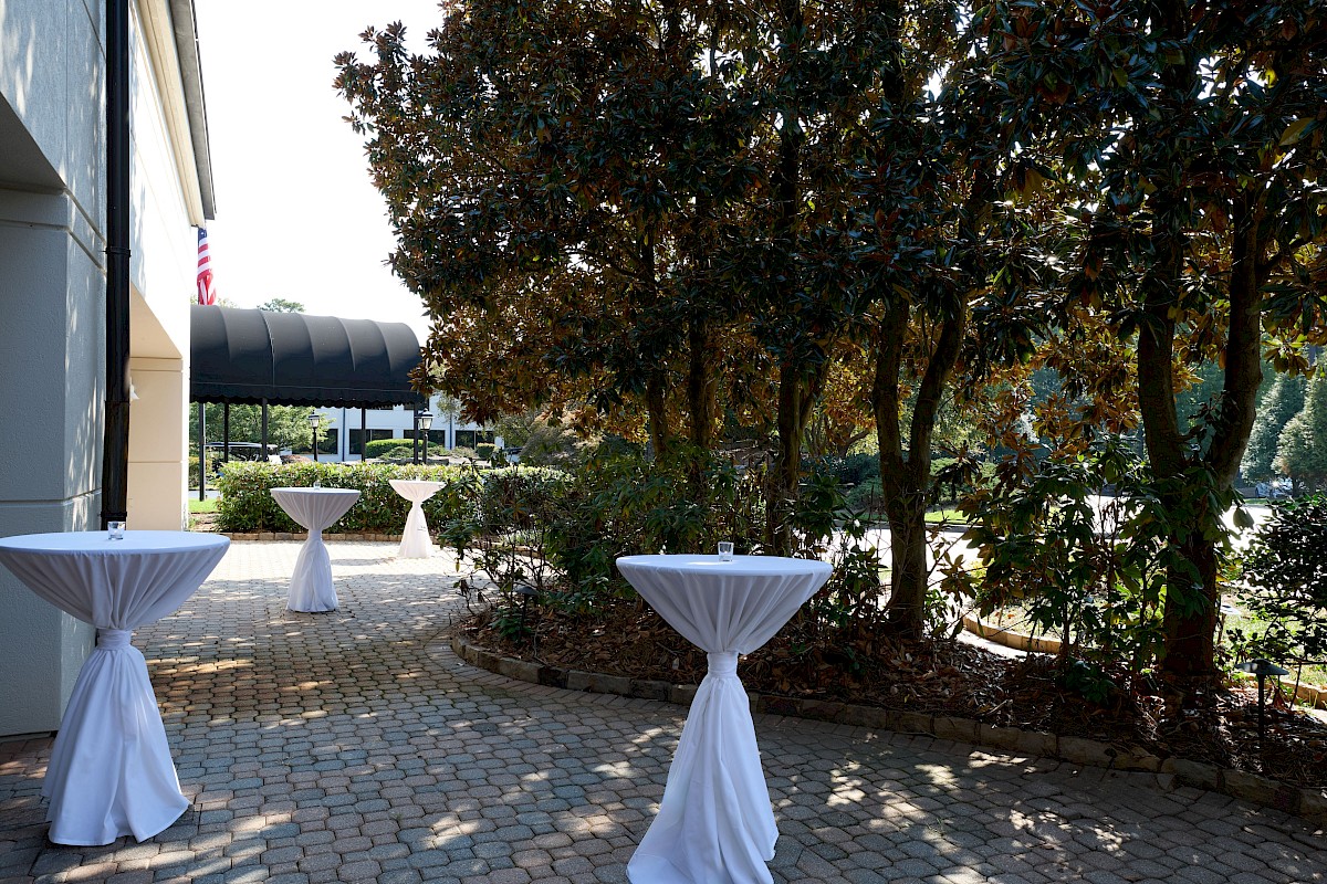 Outdoor courtyard with tree shade, several tall cocktail-height tables draped in white cloth, and paved stone ground, likely for an event.