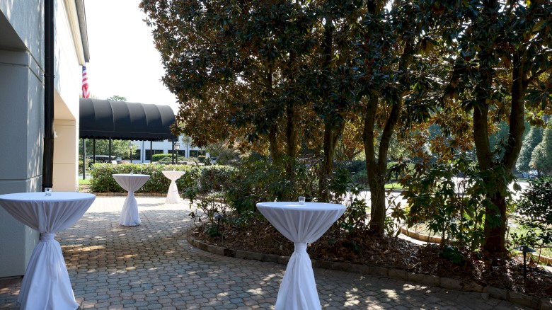 Outdoor courtyard with tree shade, several tall cocktail-height tables draped in white cloth, and paved stone ground, likely for an event.