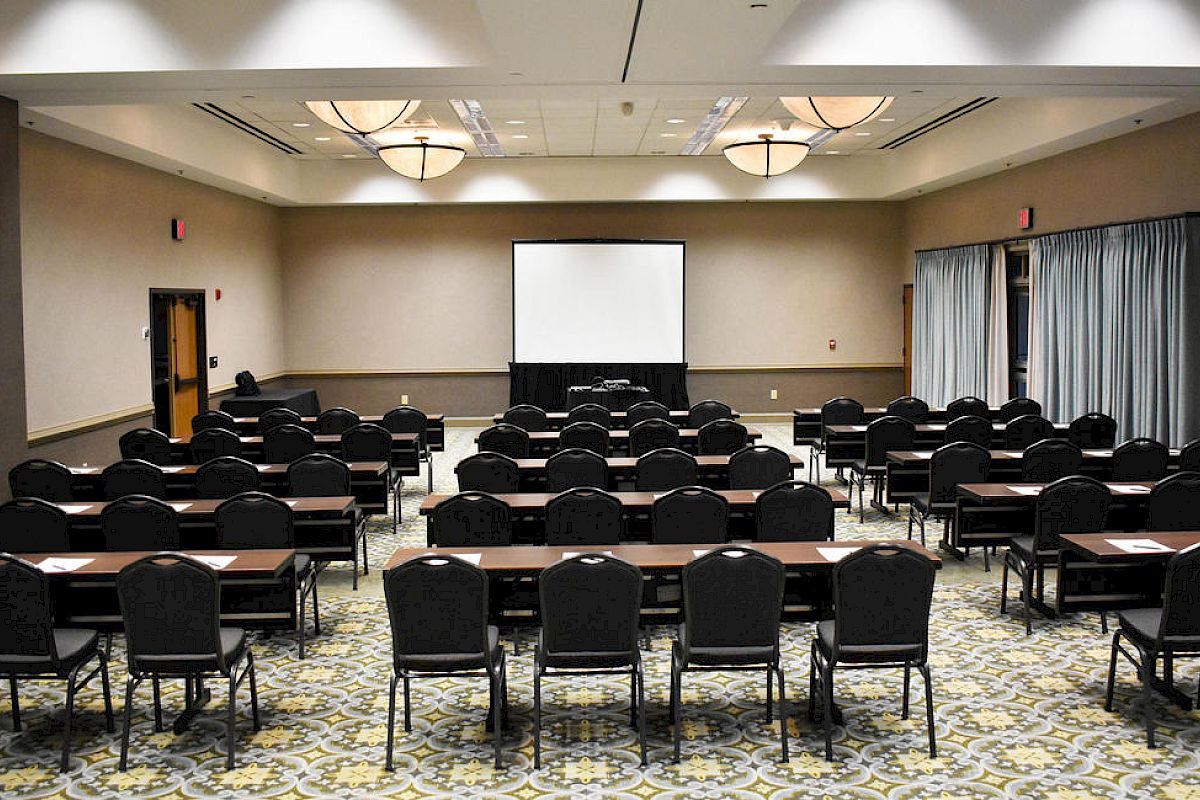 A conference room set up for a presentation with rows of chairs and tables facing a projection screen at the front, soft lighting, and a patterned carpet.