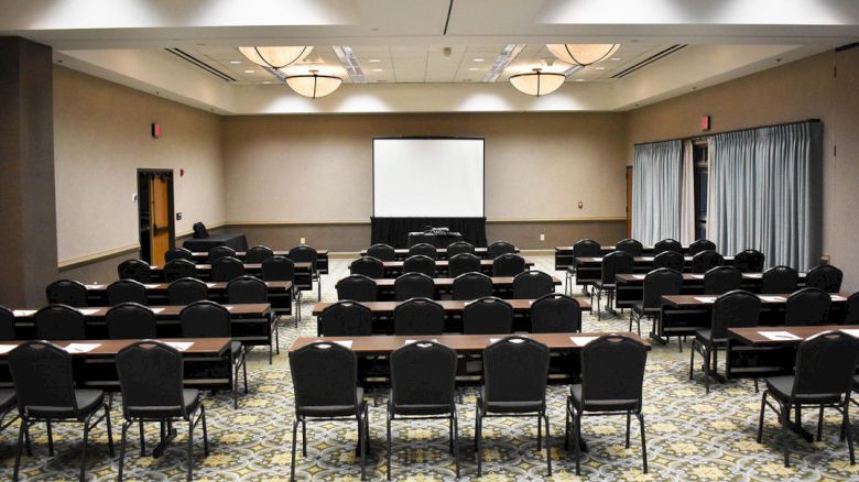 A conference room set up for a presentation with rows of chairs and tables facing a projection screen at the front, soft lighting, and a patterned carpet.