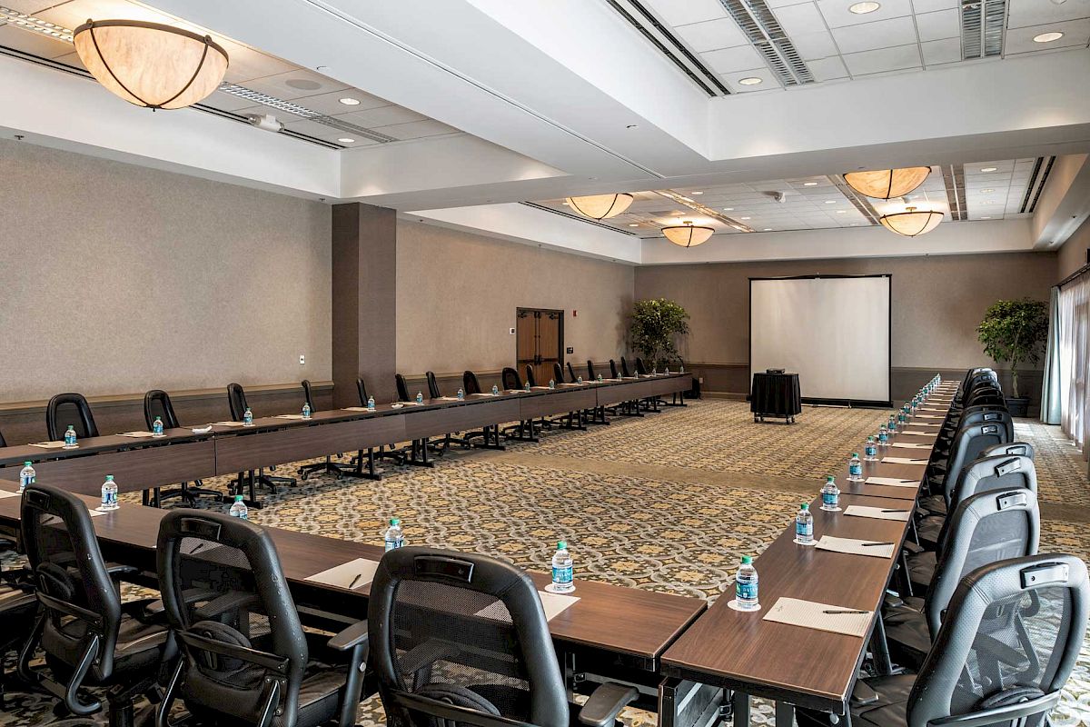A long conference room with a U/rectangular desk setup, black rolling chairs, water bottles on each desk, and a projector screen at the far end.