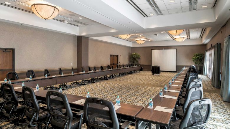 A long conference room with a U/rectangular desk setup, black rolling chairs, water bottles on each desk, and a projector screen at the far end.