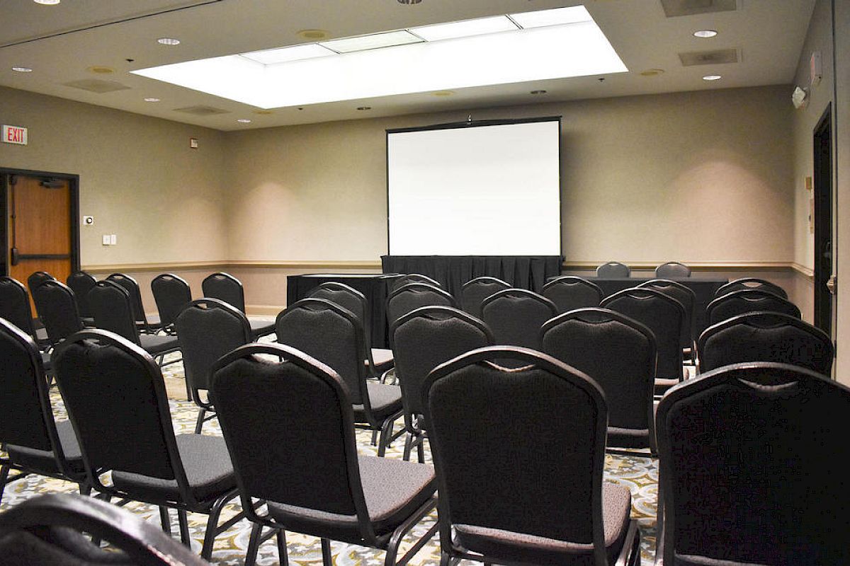 The image shows an empty conference room set up with rows of black chairs facing a projector screen at the front, with overhead lighting.