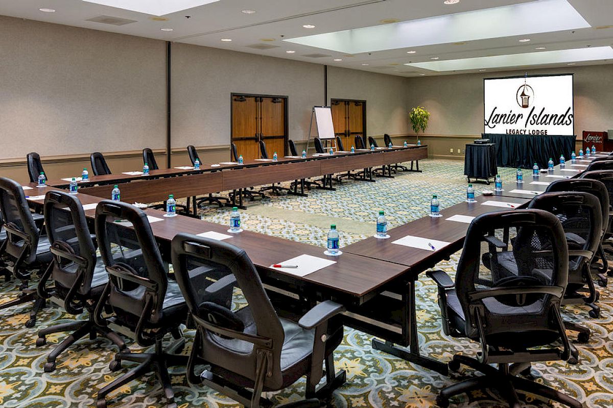 The image shows a conference room set up with a U-shaped table arrangement, chairs, water bottles, and a presentation screen displaying 