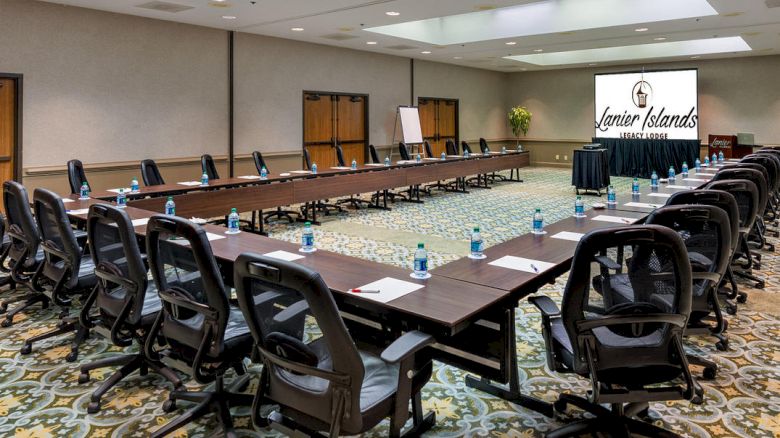 The image shows a conference room set up with a U-shaped table arrangement, chairs, water bottles, and a presentation screen displaying 