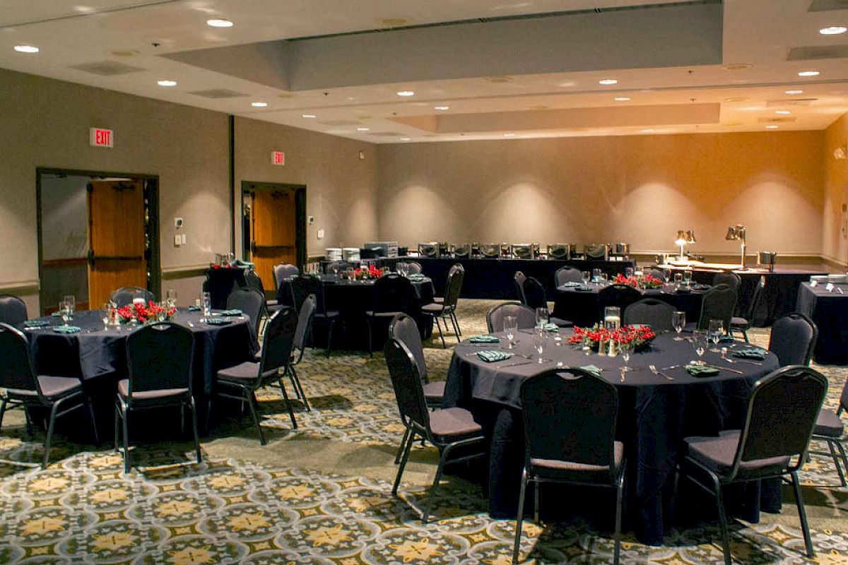 The image shows an event room set up with round tables and chairs, elegantly arranged with table settings and red centerpieces on each table.