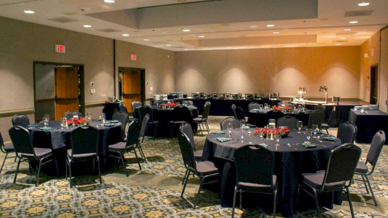 The image shows an event room set up with round tables and chairs, elegantly arranged with table settings and red centerpieces on each table.