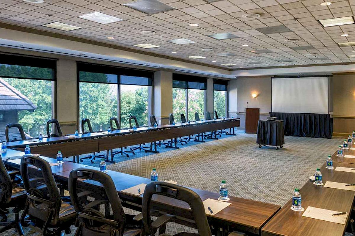 A large conference room setup with U-shaped tables, chairs, bottled water, pens, paper, and a projection screen, ready for a meeting or presentation.