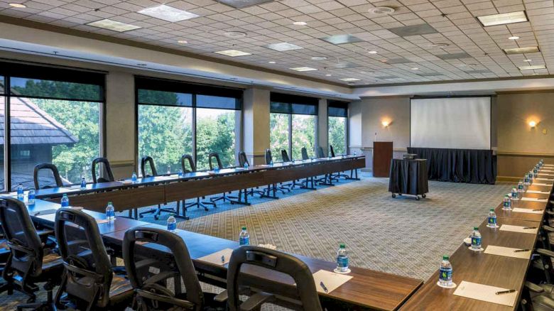 A large conference room setup with U-shaped tables, chairs, bottled water, pens, paper, and a projection screen, ready for a meeting or presentation.