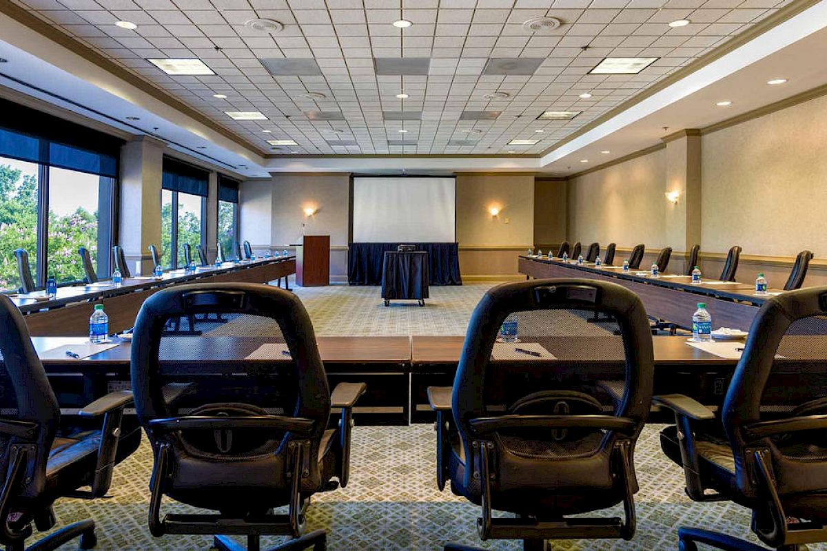 The image shows an empty conference room with long tables arranged in a U-shape, chairs, a projector screen, and water bottles on the tables.