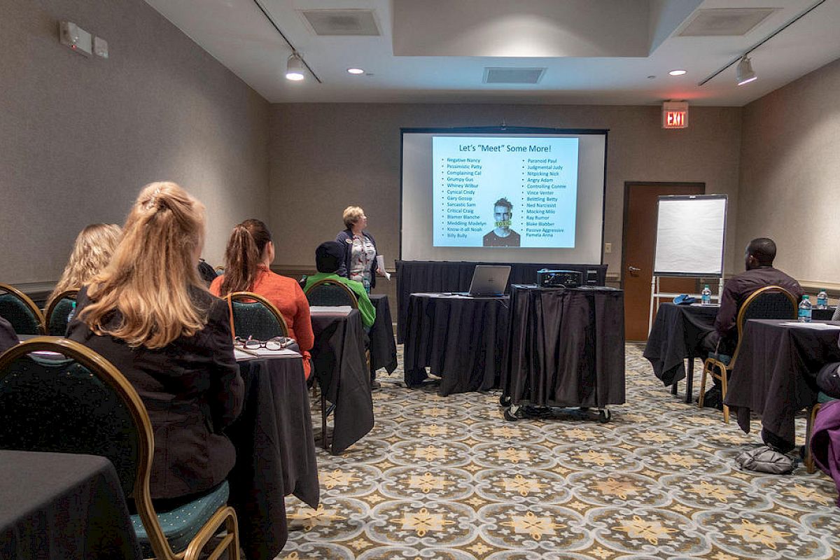 Room with people seated, a presenter at the front showing a slide with a list and picture; likely a meeting or training session.