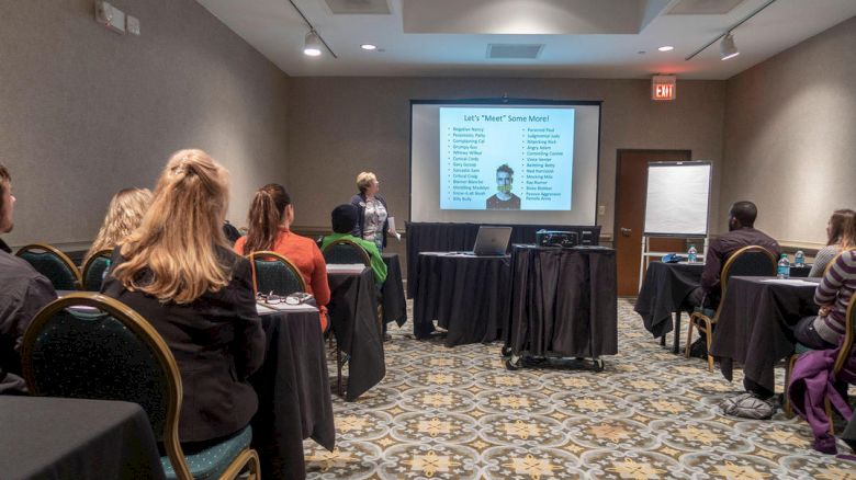 Room with people seated, a presenter at the front showing a slide with a list and picture; likely a meeting or training session.
