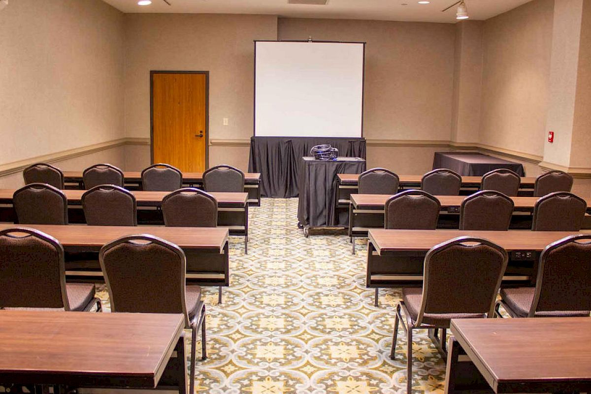 The image depicts a small conference room with rows of tables and chairs, a projector screen at the front, and a projector on a stand.