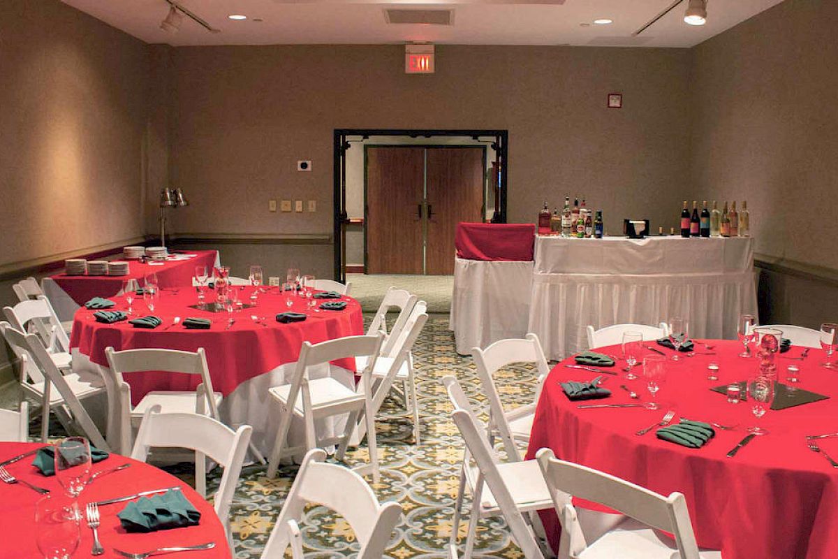 This image shows a banquet room with round tables covered in red tablecloths, white chairs, and a bar setup in the back.