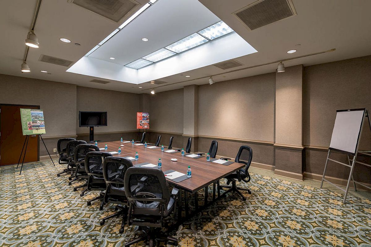This image shows a conference room with a long table, chairs, a TV, whiteboards, and posters on easels. There are water bottles and notepads on the table.