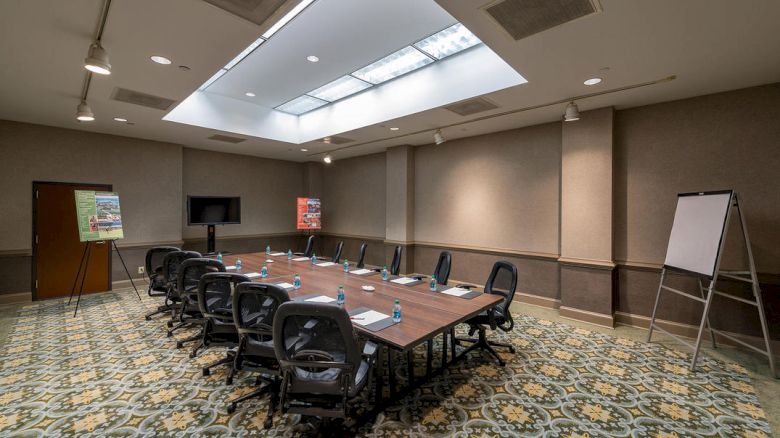 This image shows a conference room with a long table, chairs, a TV, whiteboards, and posters on easels. There are water bottles and notepads on the table.