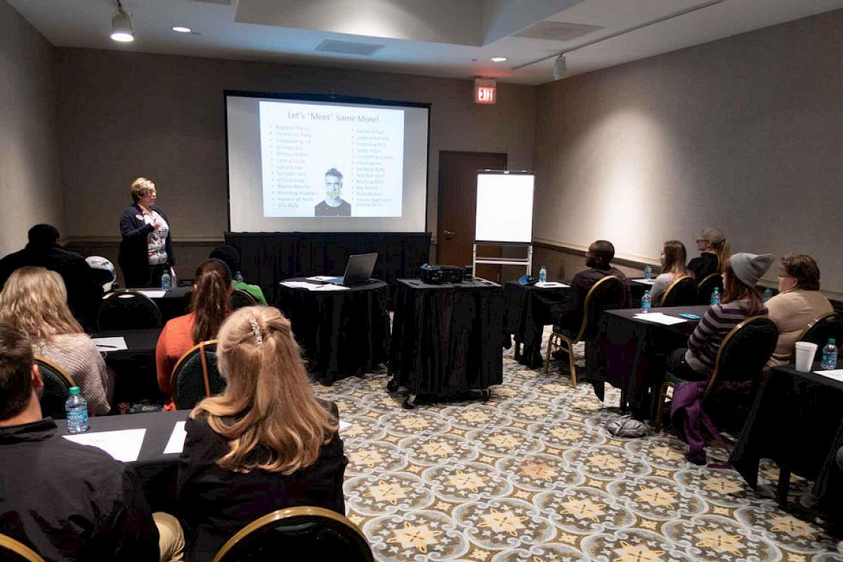 A group of people sits in a room with a patterned carpet, watching a presentation about body language. A speaker stands while explaining the content.