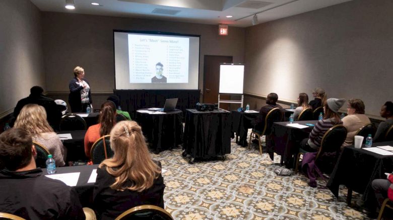 A group of people sits in a room with a patterned carpet, watching a presentation about body language. A speaker stands while explaining the content.