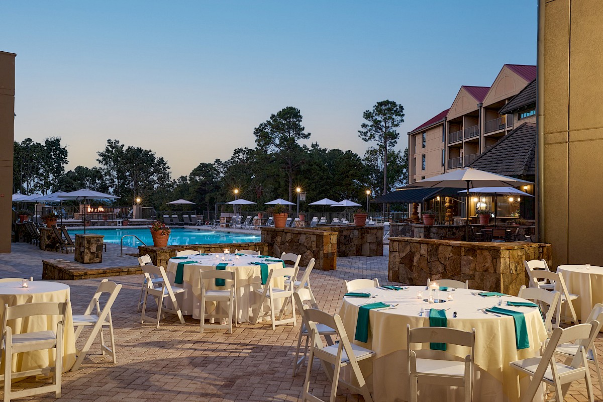Outdoor resort pool area at dusk with white plastic tables and chairs, umbrellas, stone bar, and a multi-story hotel in the background.