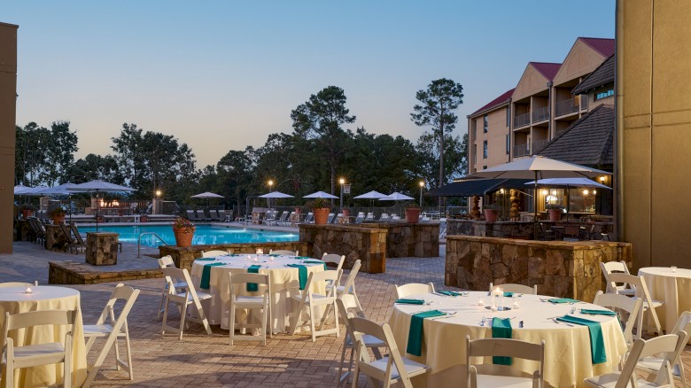 Outdoor resort pool area at dusk with white plastic tables and chairs, umbrellas, stone bar, and a multi-story hotel in the background.