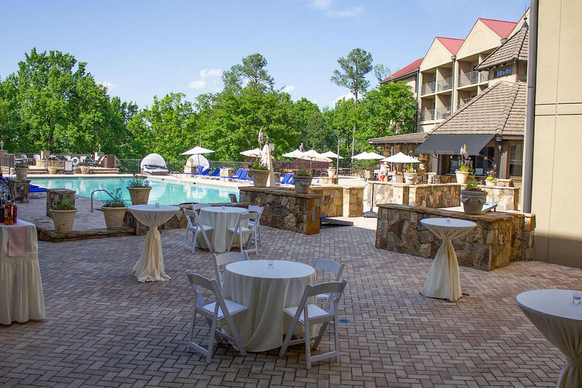 A patio area with round tables and chairs, overlooking an outdoor pool and greenery, adjacent to a multi-story building with a red roof.