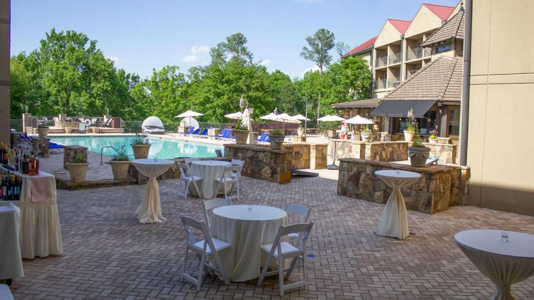A patio area with round tables and chairs, overlooking an outdoor pool and greenery, adjacent to a multi-story building with a red roof.