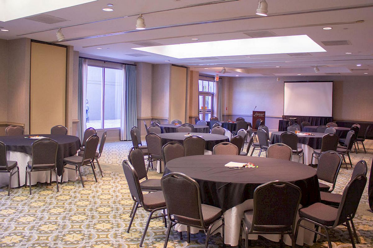 The image shows a conference room set up with round tables and chairs, a podium, and a projection screen in the background.