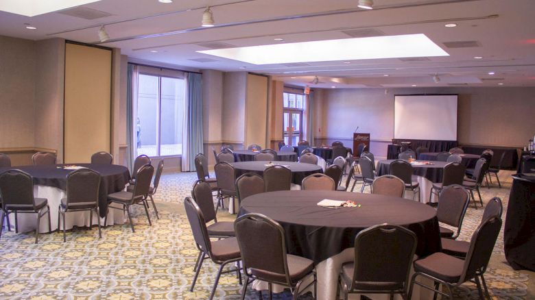 The image shows a conference room set up with round tables and chairs, a podium, and a projection screen in the background.