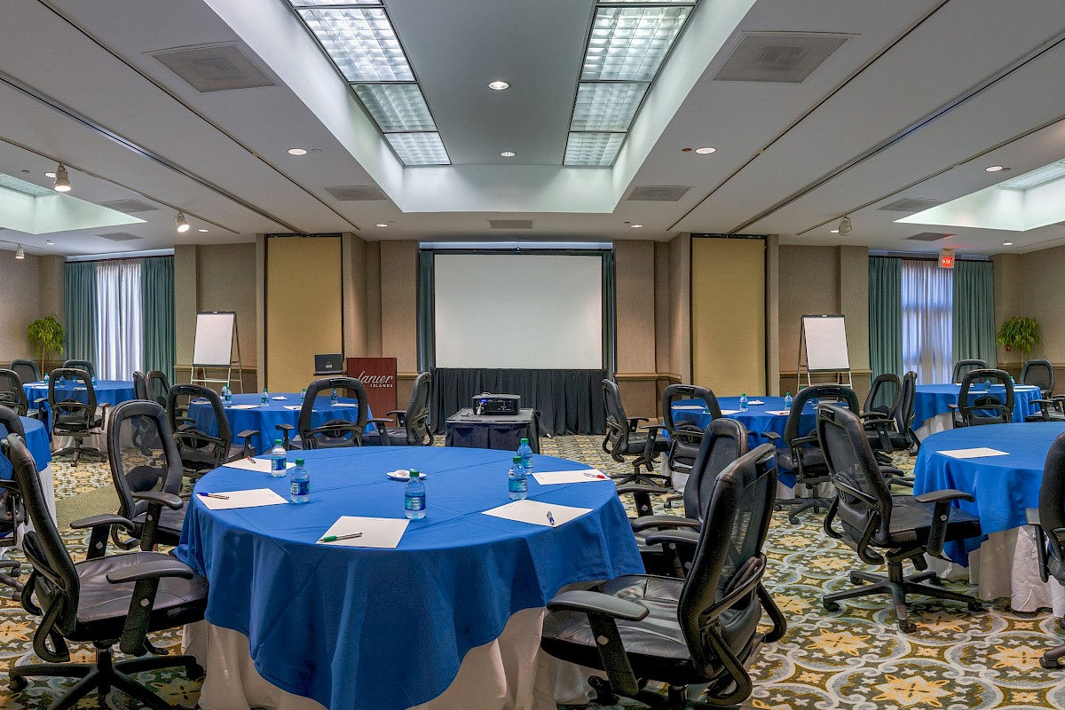 A conference room with blue round tables, black office chairs, a large projector screen, whiteboards, and ceiling lights in a spacious setup.