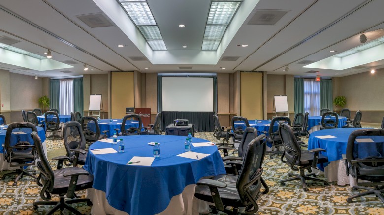 A conference room with blue round tables, black office chairs, a large projector screen, whiteboards, and ceiling lights in a spacious setup.