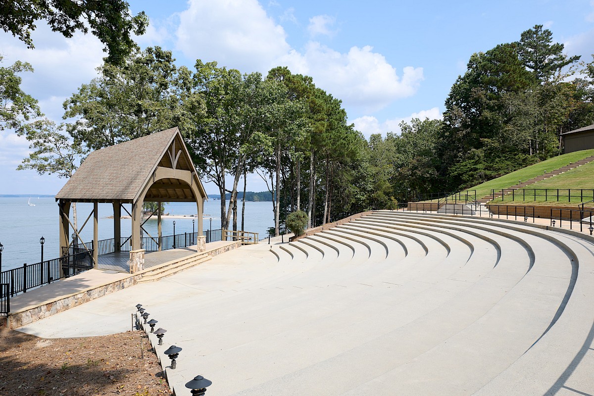 An outdoor amphitheater by a lake with curved concrete seating, a small wooden pavilion, trees, and a gentle hillside under a blue sky.