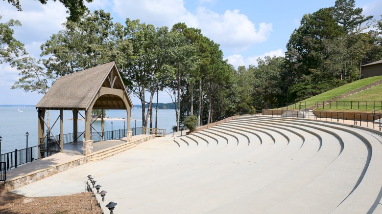 An outdoor amphitheater by a lake with curved concrete seating, a small wooden pavilion, trees, and a gentle hillside under a blue sky.