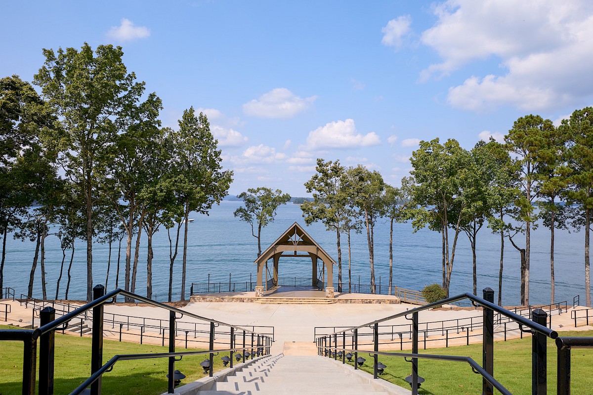 A scenic lakeside park with a central stone staircase leading to a wooden gazebo, framed by tall trees and calm water in the background.