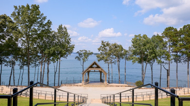 A scenic lakeside park with a central stone staircase leading to a wooden gazebo, framed by tall trees and calm water in the background.