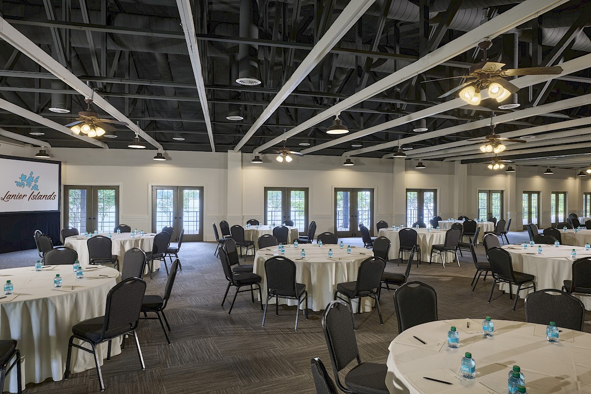 A spacious event hall with round tables, black chairs, table decorations, and ceiling fans, set up for a conference or banquet.
