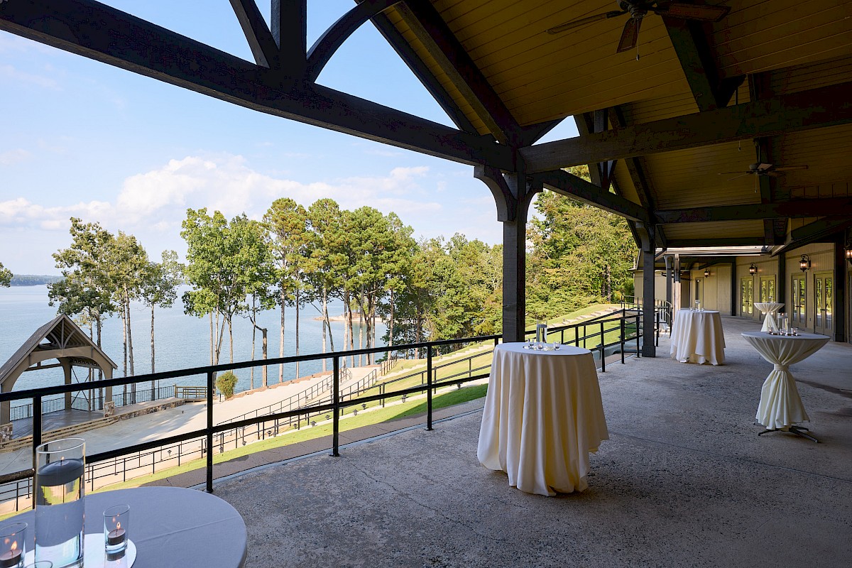 Outdoor event area with high ceilings, string lights, and tall cocktail tables on a balcony overlooking a lake and trees, perfect for a reception.