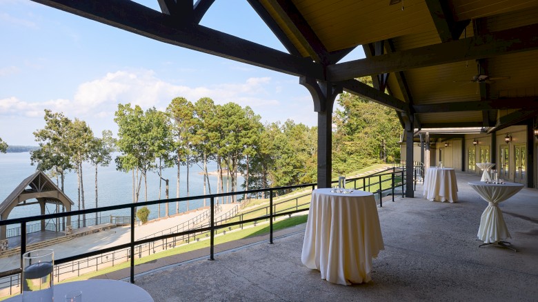 Outdoor event area with high ceilings, string lights, and tall cocktail tables on a balcony overlooking a lake and trees, perfect for a reception.