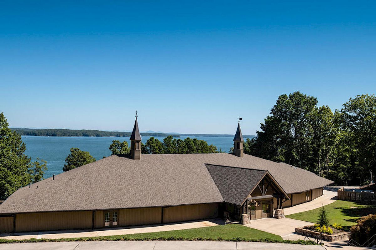 A large lodge by a lake with a grassy yard, brown roof, twin small towers, surrounding trees, and clear blue sky, inviting and serene.