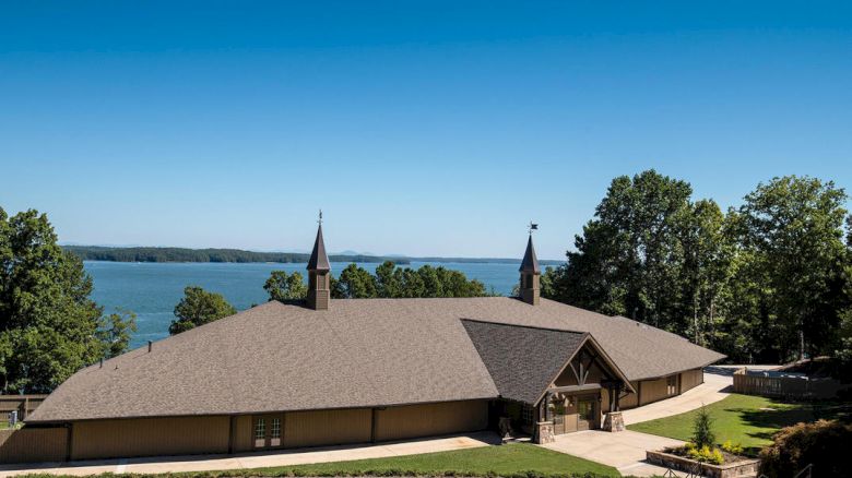 A large lodge by a lake with a grassy yard, brown roof, twin small towers, surrounding trees, and clear blue sky, inviting and serene.
