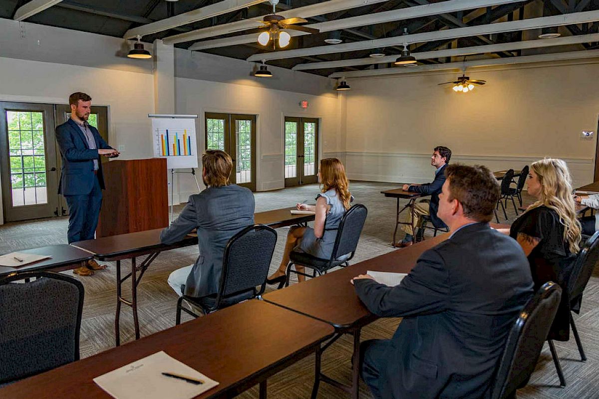 A man presents at a podium to a small audience in a conference room, with charts on a stand and attendees seated at tables.