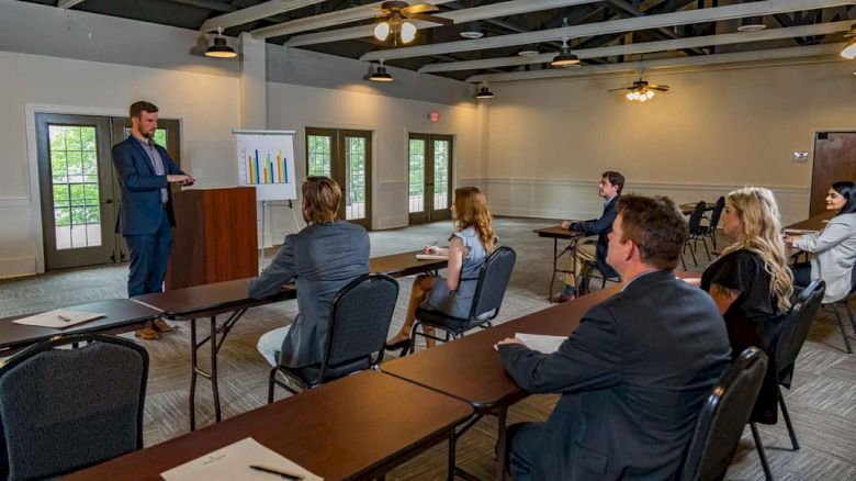 A man presents at a podium to a small audience in a conference room, with charts on a stand and attendees seated at tables.