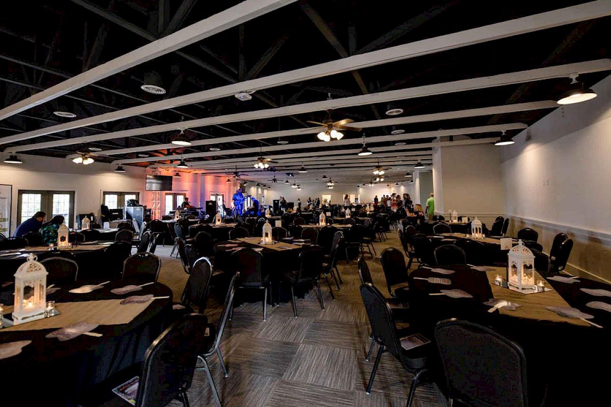 A large conference or banquet hall with round and rectangular tables, chairs, and lantern centerpieces; people gather toward the back, dim lighting.