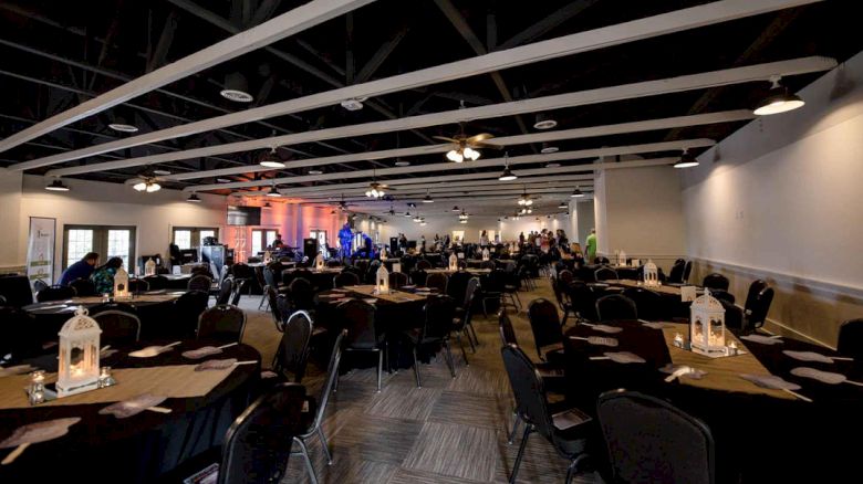 A large conference or banquet hall with round and rectangular tables, chairs, and lantern centerpieces; people gather toward the back, dim lighting.