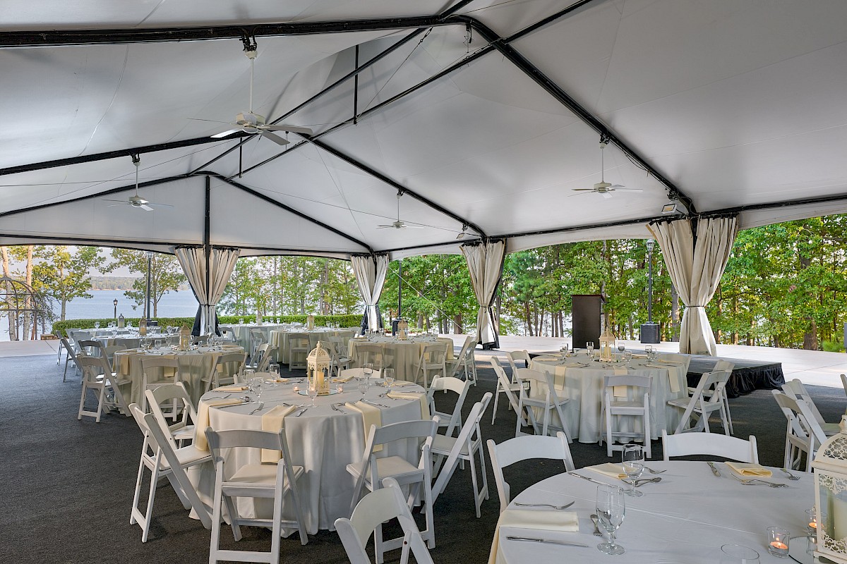 Outdoor event setup under a white tent with round tables, white linens, and folding chairs, ready for a formal reception on a sunny day.