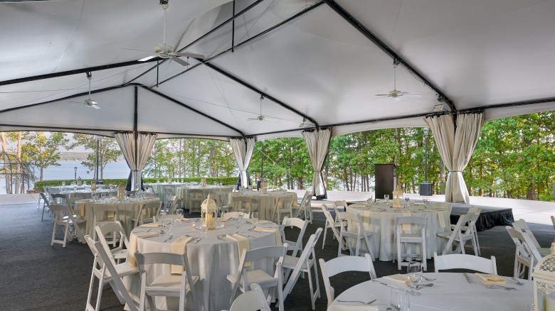 Outdoor event setup under a white tent with round tables, white linens, and folding chairs, ready for a formal reception on a sunny day.